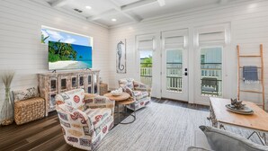 A spacious room with white walls, a ceiling fan, a large TV, a wooden table, and a view of the outdoors through a sliding glass door. - Private Pool, Golf Cart Included, Boardwalk to Beach (Port Aransas)