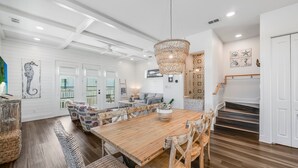 A spacious dining area with a wooden table and wicker chairs, a chandelier, and a staircase leading to a second floor. - Private Pool, Golf Cart Included, Boardwalk to Beach (Port Aransas)