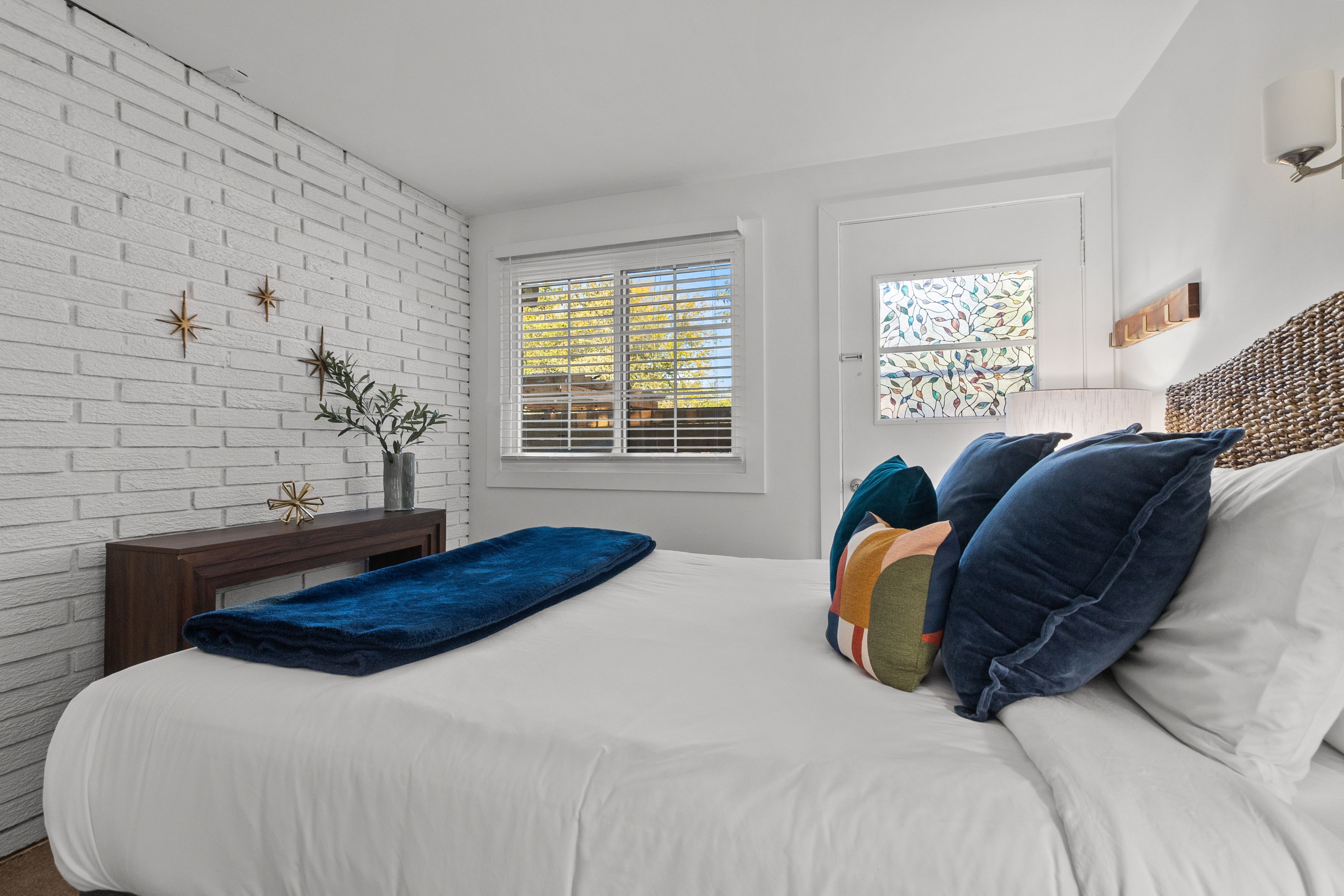 Un dormitorio con pared de ladrillo blanco, una ventana con persianas, cama con ropa de cama blanca y varias almohadas, mesita de noche con un florero y un adorno en la pared.