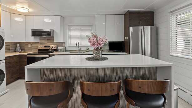 Une cuisine moderne avec des meubles blancs, un îlot central doté de chaises, un four à micro-ondes, une cuisinière et un réfrigérateur.