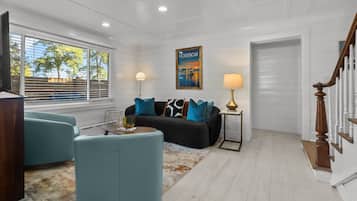 Ein Wohnzimmer mit einem schwarzen Sofa, blauen Sesseln, einem Couchtisch und einem Teppich. An der Wand hängt ein Gemälde und zur Rechten führt eine Treppe hinauf.