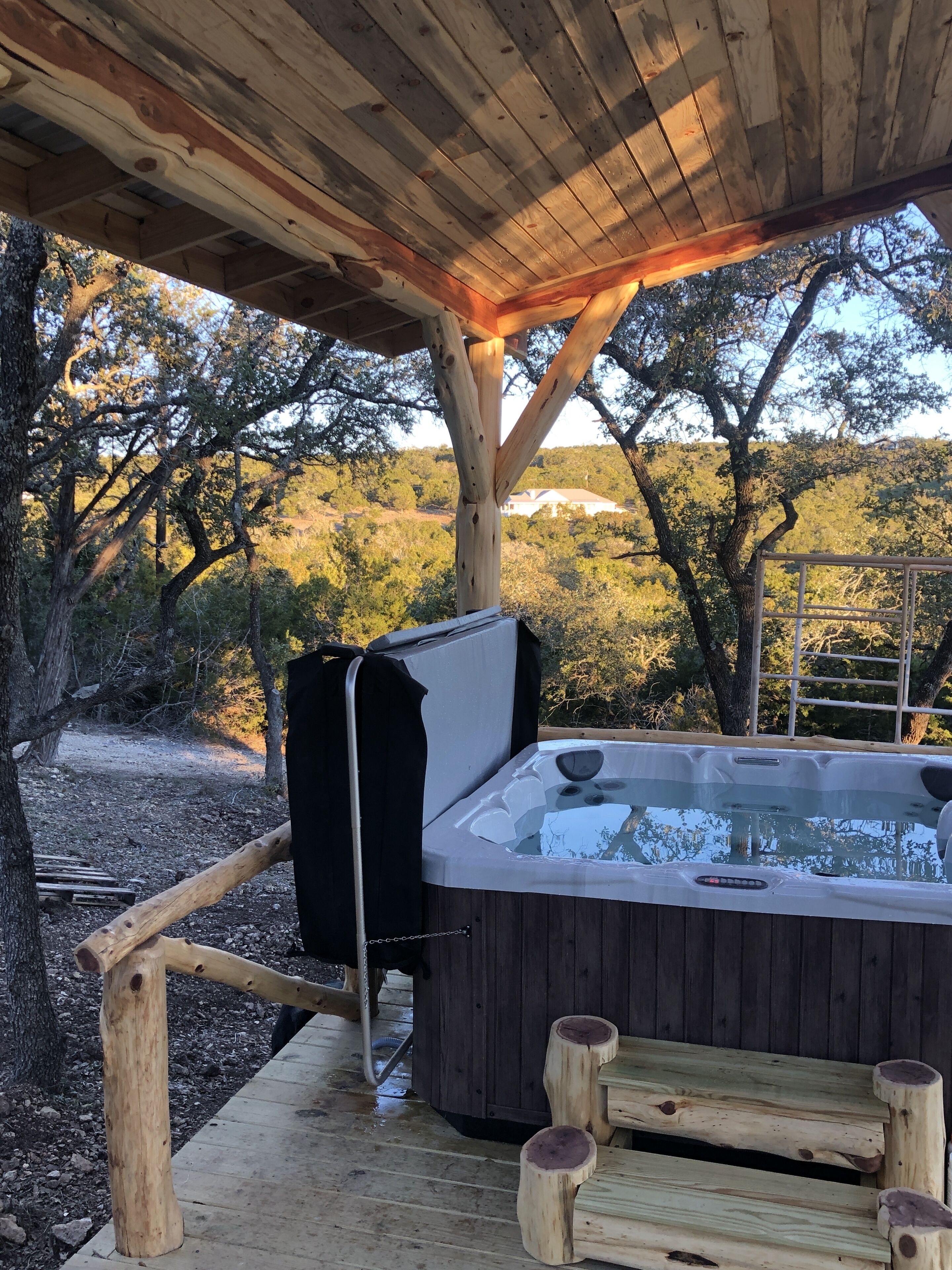 Un jacuzzi bajo una pérgola de madera con vistas a árboles y un edificio lejano.