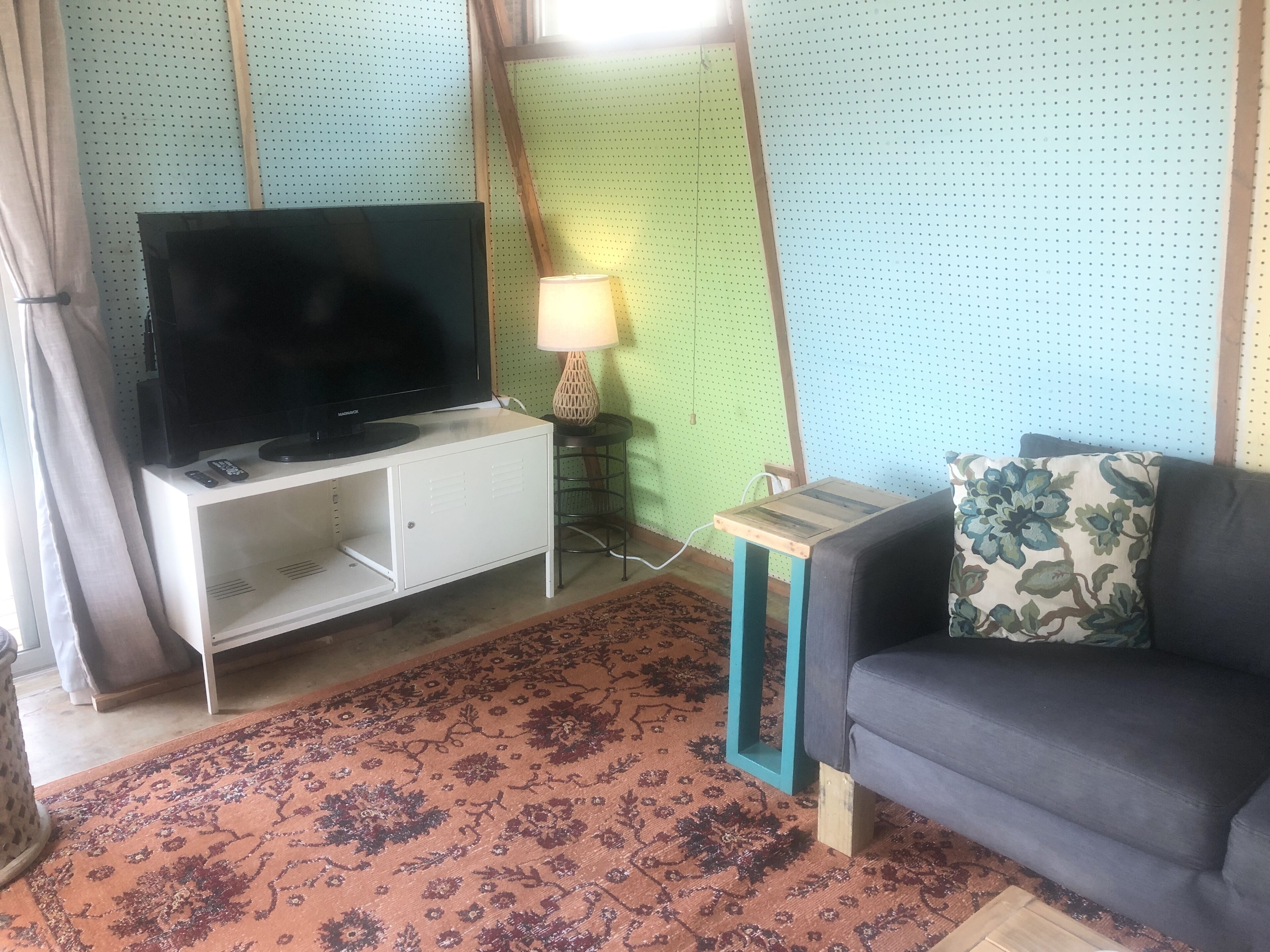 Una sala de estar con una televisión sobre un mueble blanco, una mesita con una lámpara, un sofá con un cojín floral y una alfombra estampada.