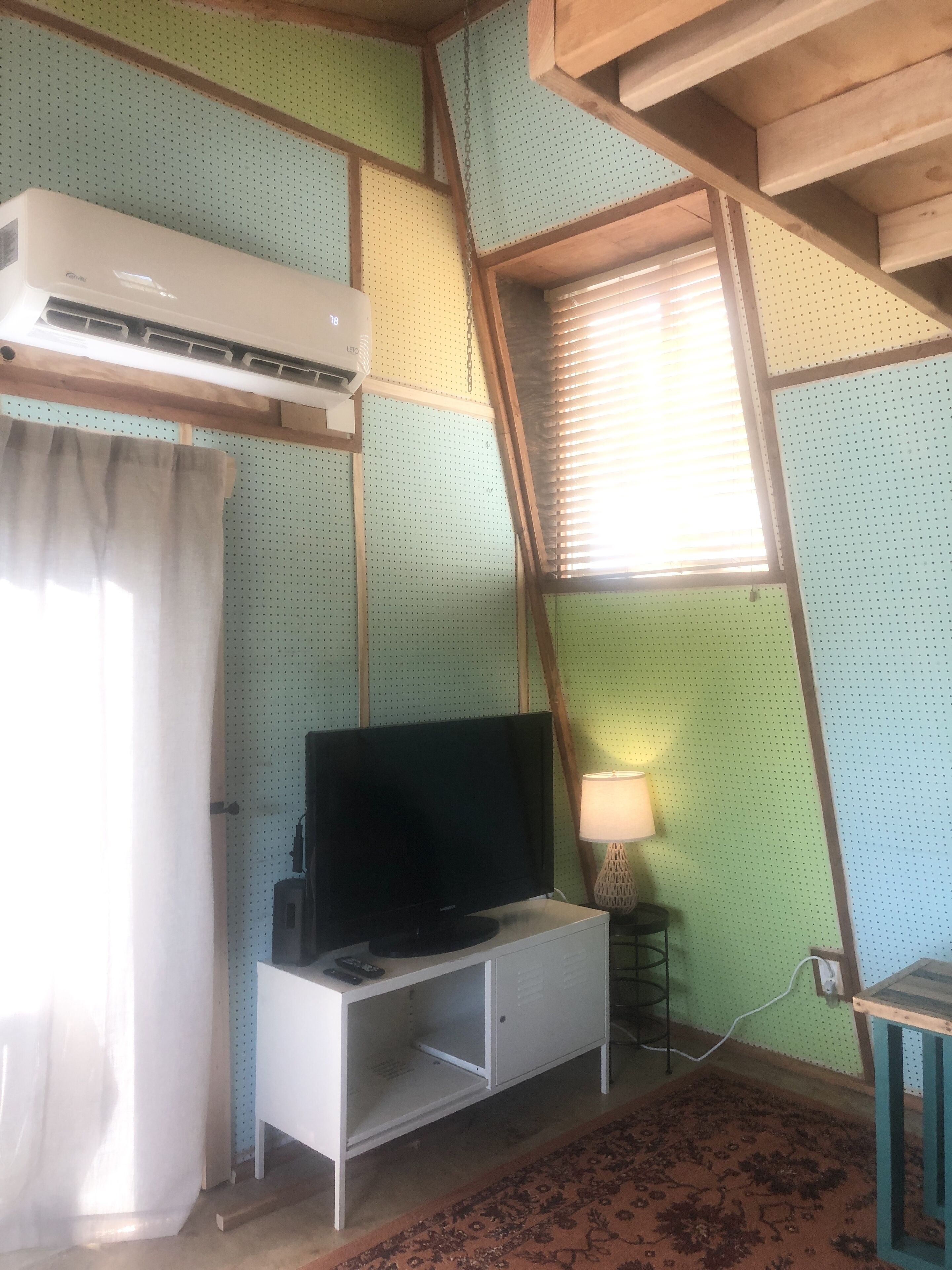 Ein Zimmer mit Fernseher auf einer weißen Kommode, einer Wandklimaanlage, einer Lampe auf einem kleinen Tisch und einem gemusterten Teppich.