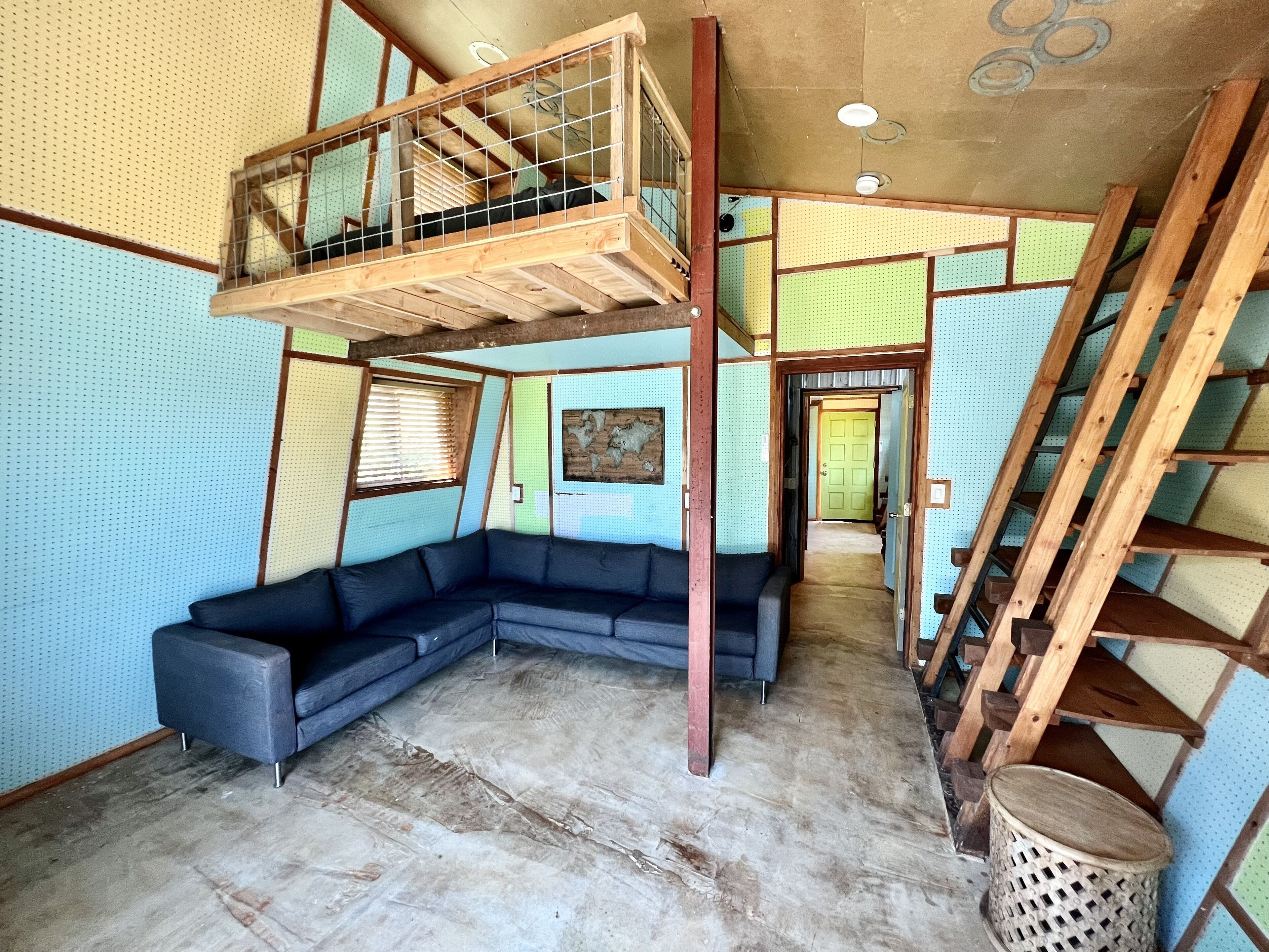 A room with a sectional sofa, a wooden loft with a metal railing, a ladder, and a woven basket.