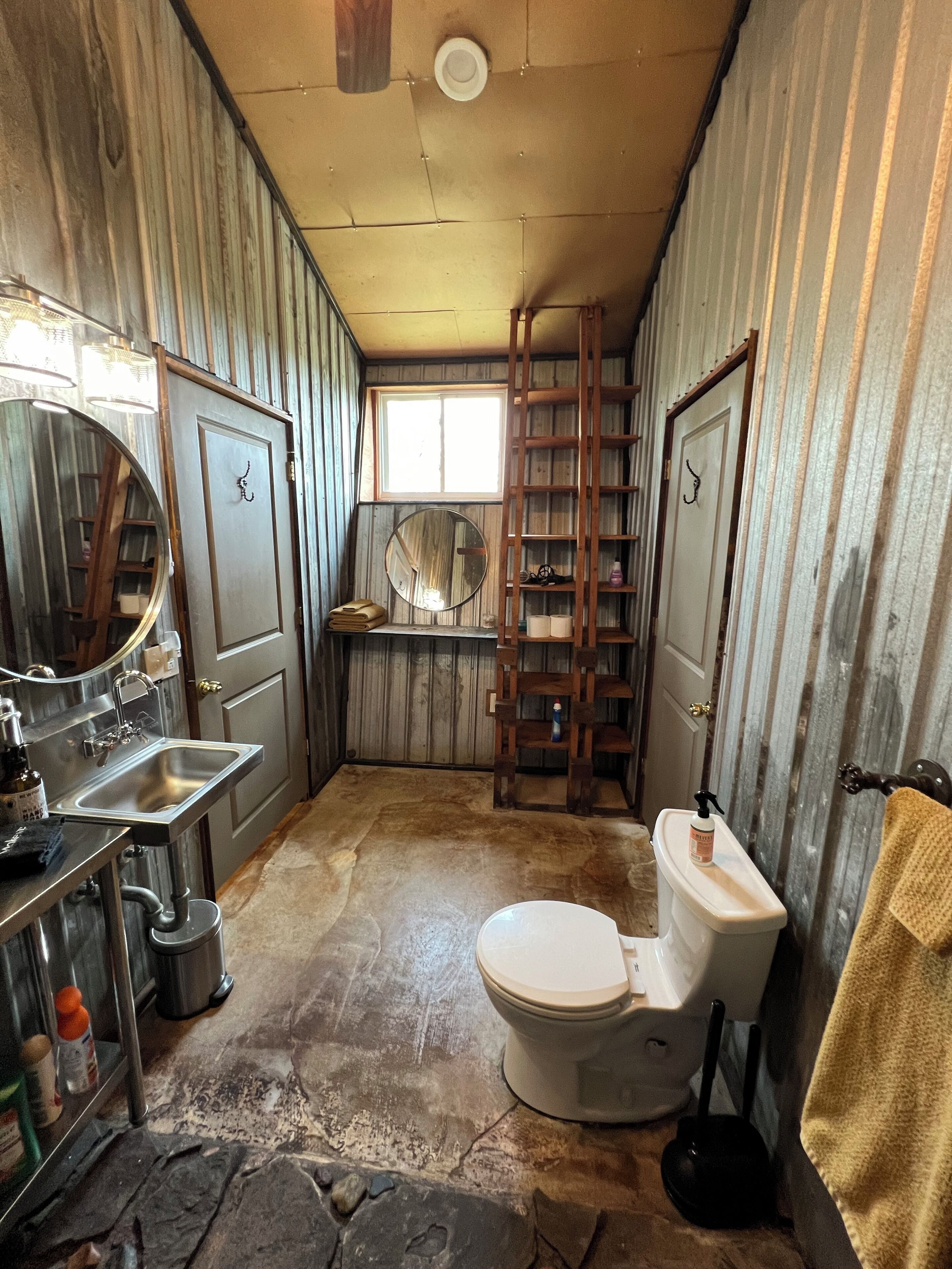 Une salle de bain rustique avec un sol en pierre, un lavabo avec un miroir, des toilettes et une échelle en bois.