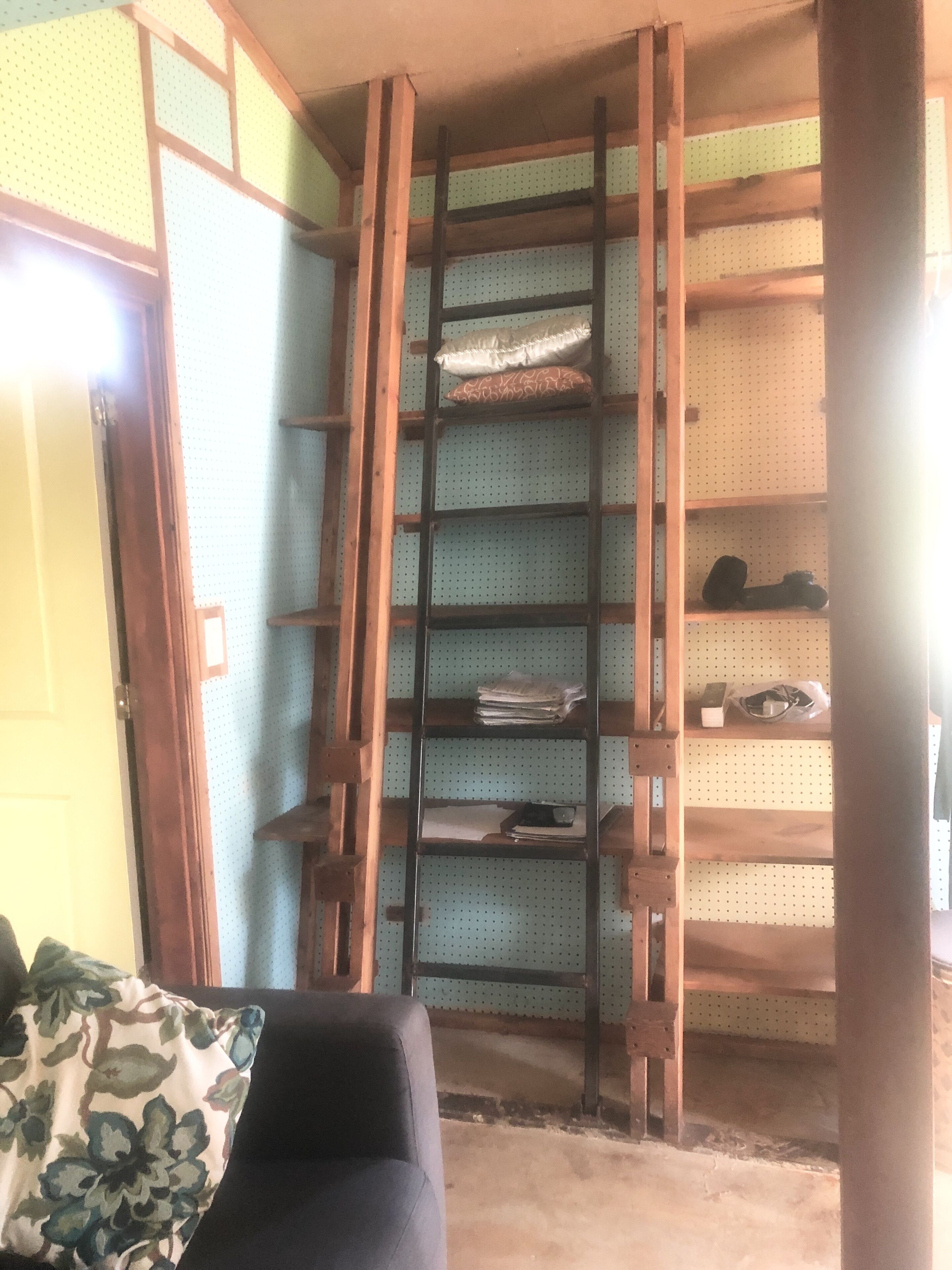 A room with a wooden ladder leading to shelves, a door, and a patterned pillow on a couch.