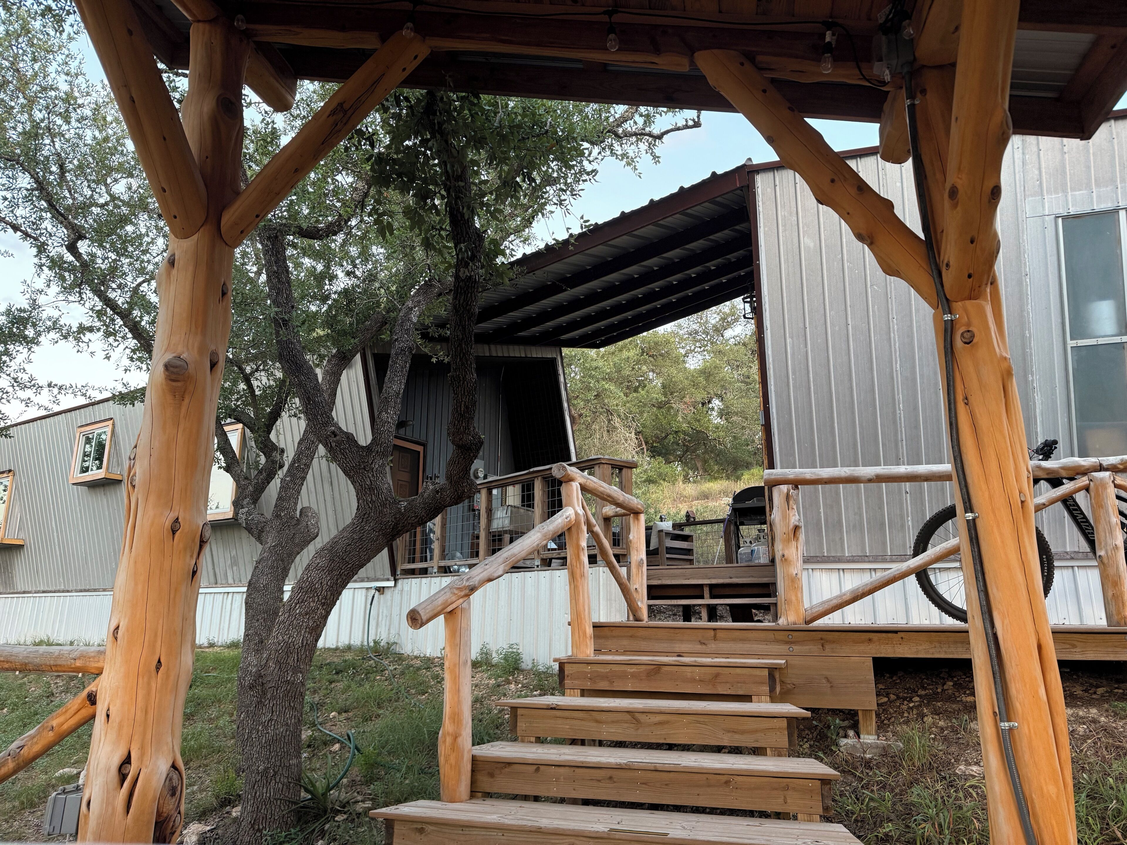 Eine Holztreppe führt zu einer Terrasse mit Metallgeländer, umgeben von Bäumen und einem Gebäude mit Wellblechfassade.