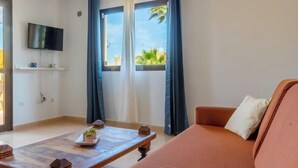 A living room with a sofa, a coffee table with decorative items, a flat-screen TV mounted on the wall, and a window with curtains. - Cueva Azul Corralejo by iRent Fuerteventura (Corralejo)