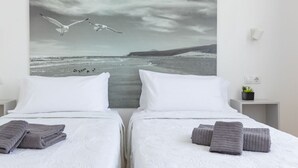 A hotel room with two beds, white bedding, and a large seascape painting above the beds. - Cueva Azul Corralejo by iRent Fuerteventura (Corralejo)