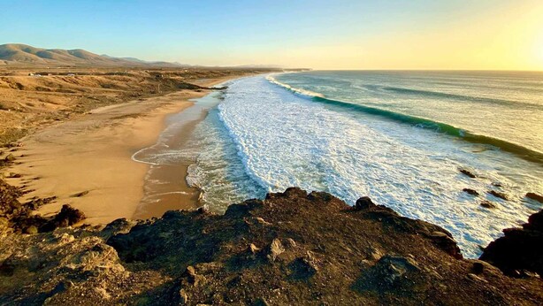 A coastal landscape with a sandy beach, rocky shoreline, and distant mountains under a clear sky. - Cueva Azul Corralejo by iRent Fuerteventura (Corralejo)
