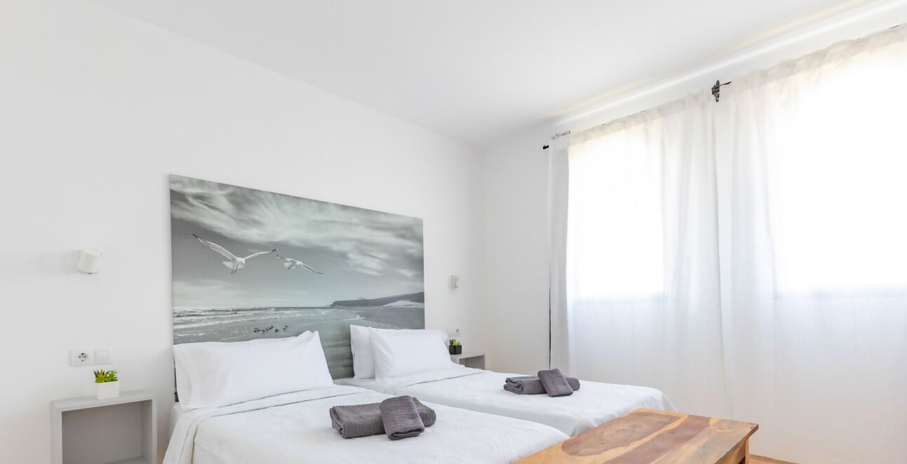 A hotel room with two beds, a wooden table, a painting of seagulls, and a view of the sea.
