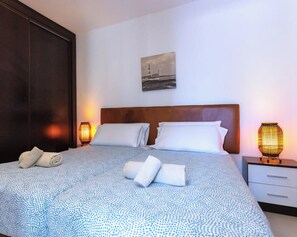 Una cama bien hecha con una colcha estampada, dos almohadas y toallas enrolladas sobre una pared blanca con un cuadro colgado.