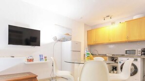 A compact living space with a flat-screen TV mounted on the wall, a small dining area with a glass table, white chairs, a white washing machine, and a microwave.