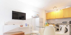 Un espacio habitable compacto con un televisor de pantalla plana montado en la pared, una pequeña zona de comedor con una mesa de vidrio, sillas blancas, una lavadora blanca y un microondas.