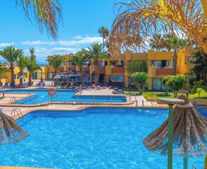 Zona de piscina de hotel con varias piscinas, sillas de descanso y palmeras.