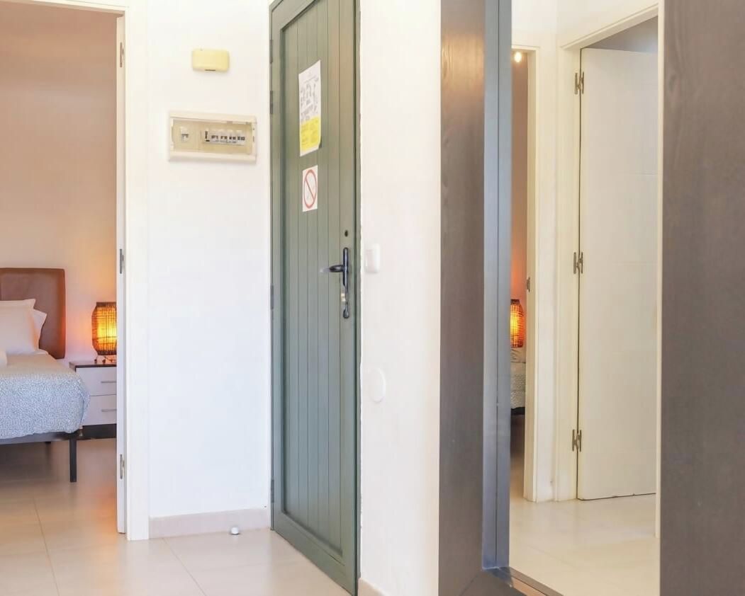 Una habitación de hotel con cama, mesita de noche, lámpara y una puerta que da a otra estancia.