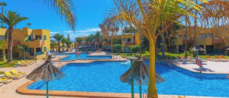 Zona de piscina en un resort, con varias piscinas, tumbonas y palmeras.