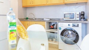 A compact kitchen with wooden cabinets, a microwave, a washing machine, and a glass table.