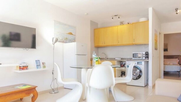 A modern kitchen with a microwave, oven, and washing machine. There is a dining area with white chairs and a table.