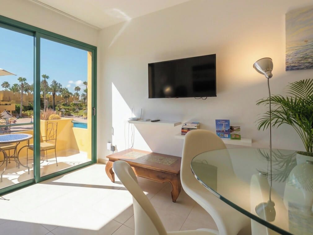 A modern living room with a glass table, a flat-screen TV mounted on the wall, and a view of the outdoor pool area.