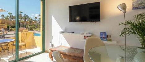 A modern living room with a glass table, a flat-screen TV mounted on the wall, and a view of the outdoor pool area.