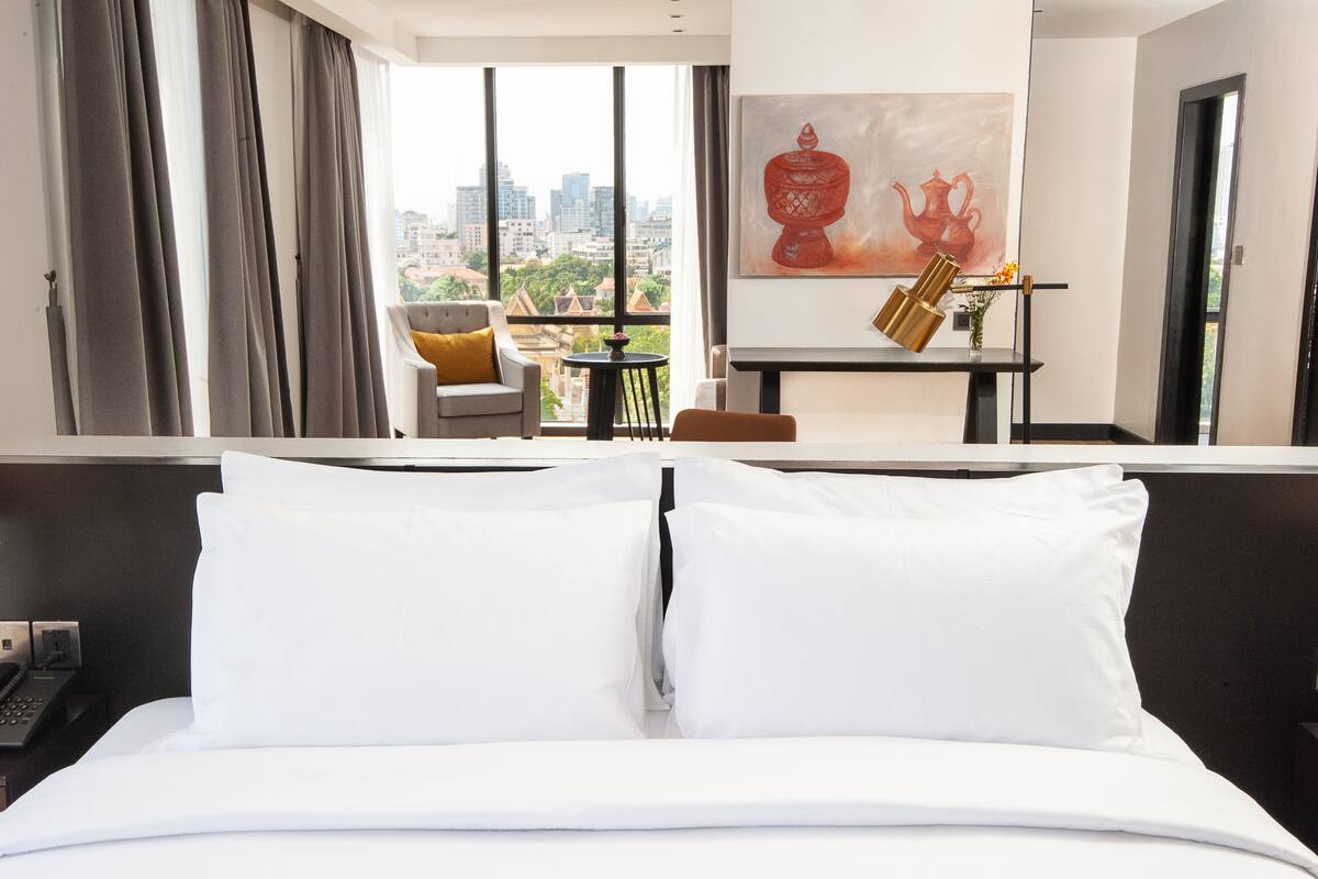 Une chambre d’hôtel avec un grand lit, une vue sur la ville, un tableau au mur et une petite table sur laquelle est posé un vase.