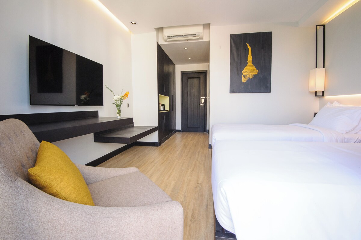 Een moderne hotelkamer met een bed, een bank met een geel kussen, een tv aan de muur, een kunstwerk en een trap naar een andere kamer.