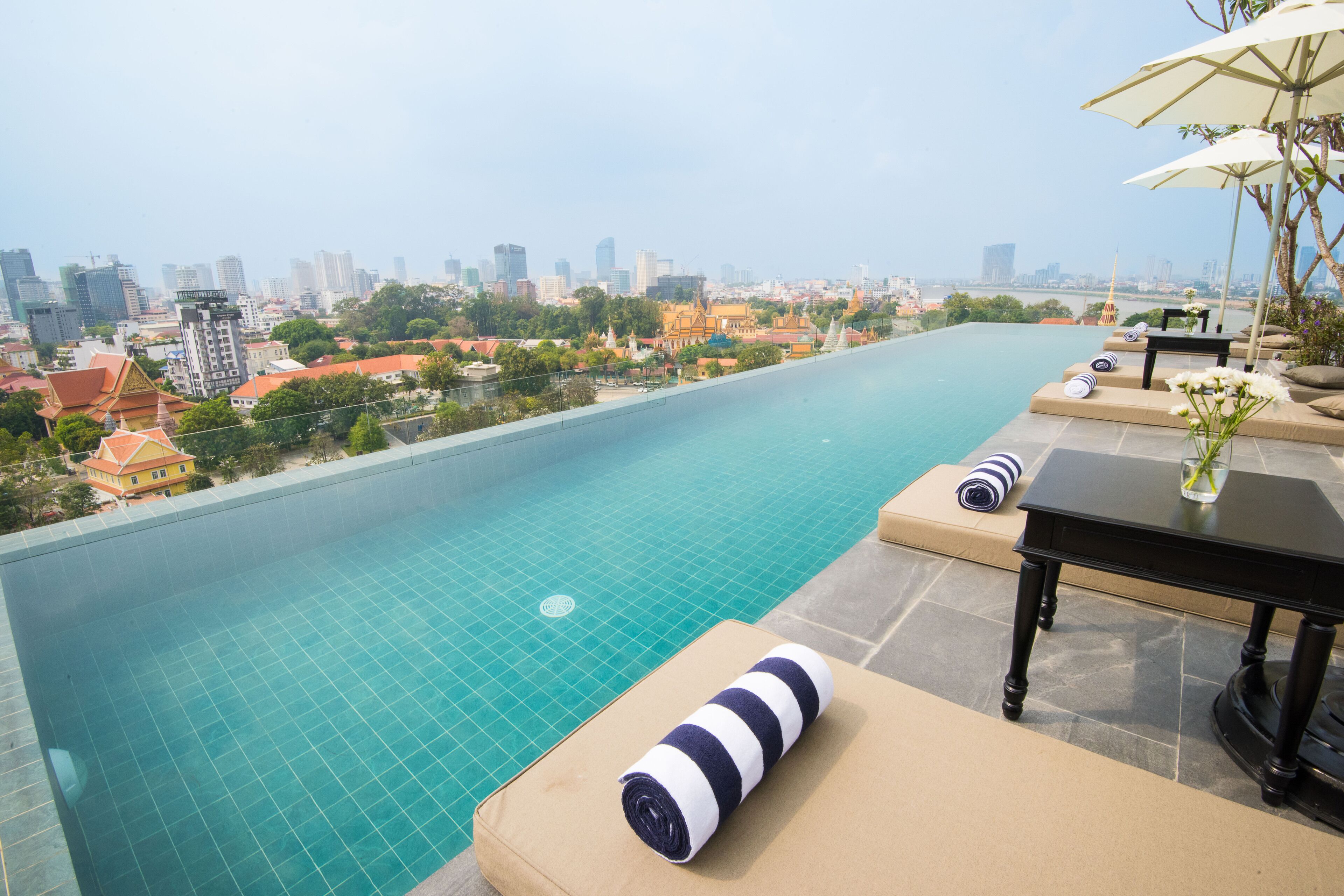 Une piscine à débordement située sur le toit, offrant une vue sur la ville, un espace détente avec des coussins et une table avec un parasol.