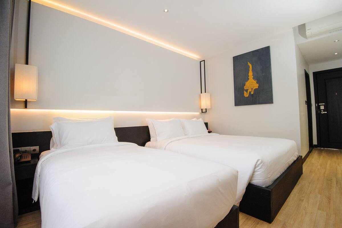 Une chambre d’hôtel avec deux lits, une table de chevet avec un téléphone, une lampe fixée au mur et un tableau représentant un temple.