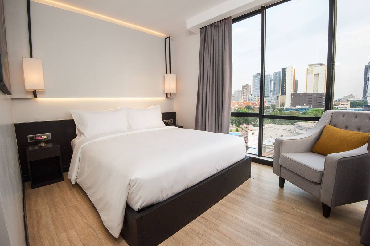 Une chambre d’hôtel moderne avec un grand lit, un fauteuil gris et une vue sur la ville à travers de grandes fenêtres.