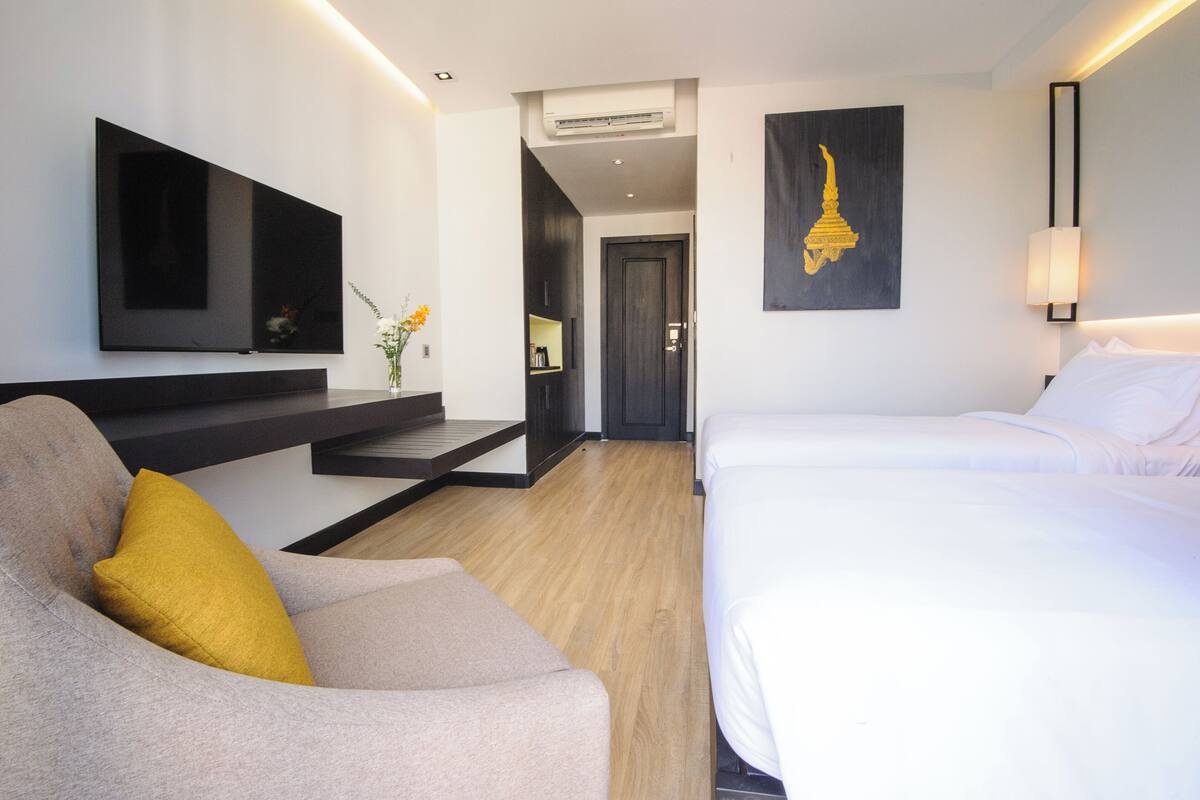 Une chambre d’hôtel moderne comprenant un lit, un canapé avec un coussin jaune, un téléviseur fixé au mur, une œuvre d’art accrochée au mur et un escalier menant à une autre pièce.