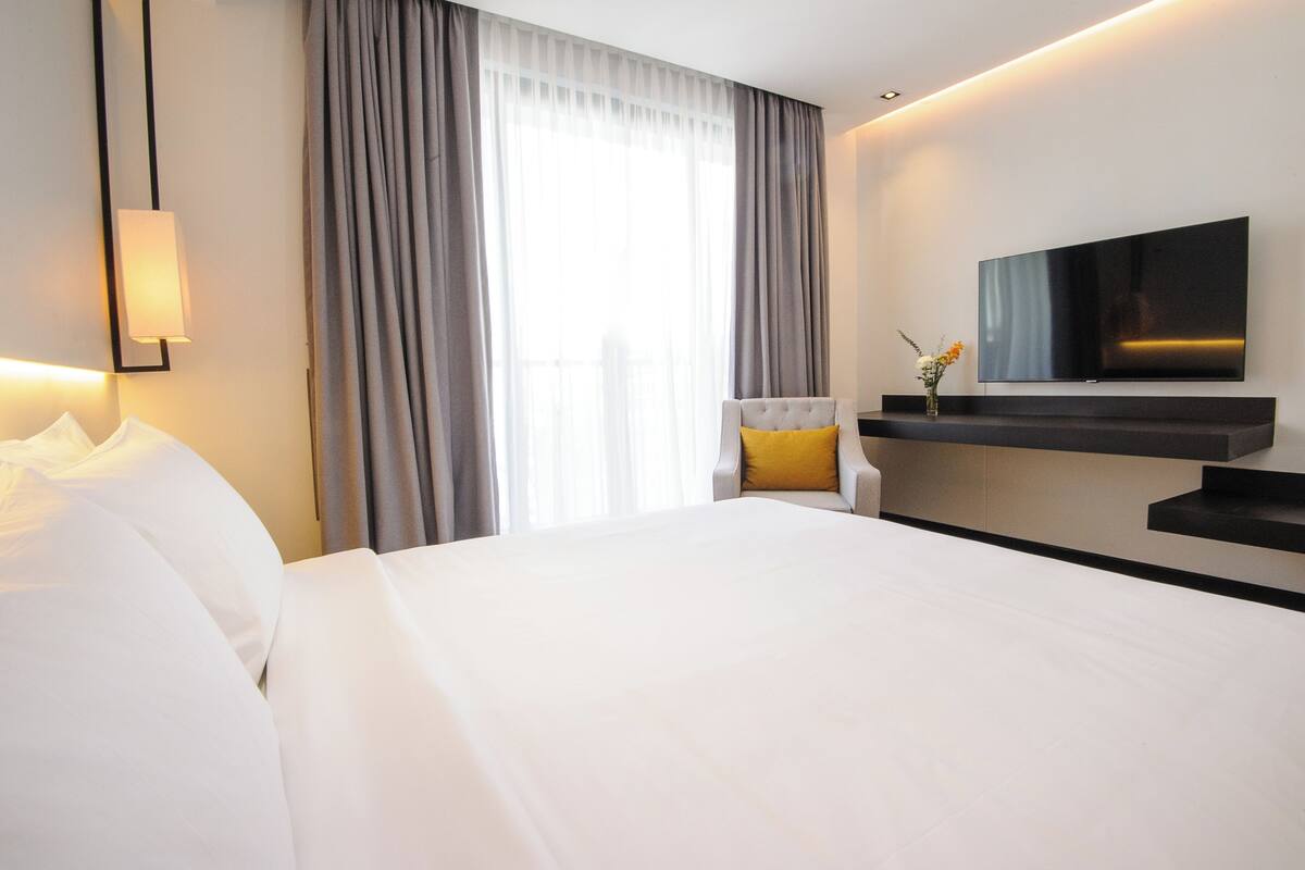 Une chambre d’hôtel avec un grand lit, une chaise avec un coussin jaune, un téléviseur à écran plat fixé au mur et une fenêtre avec des rideaux.