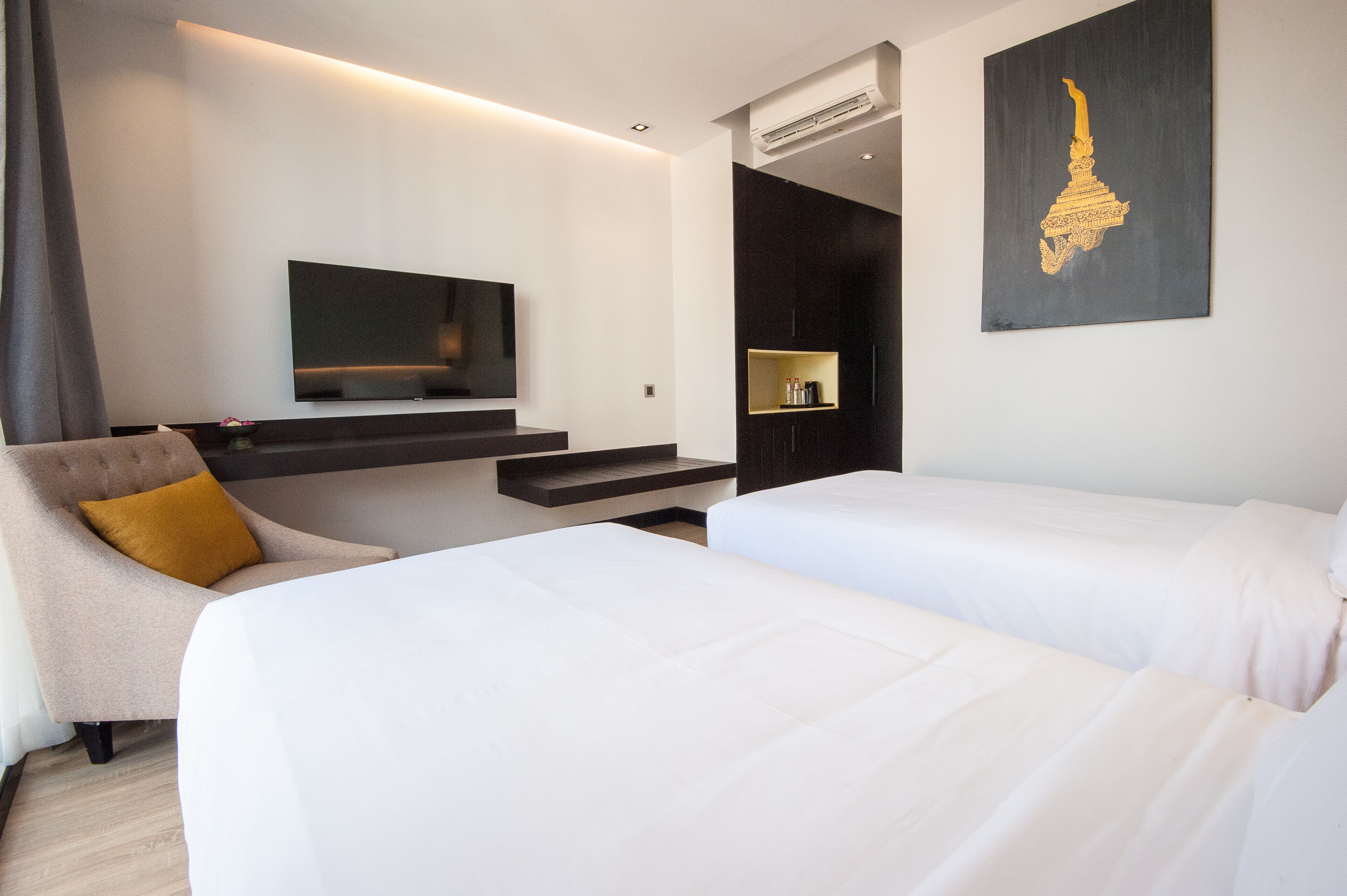 Een moderne hotelkamer met een flatscreen televisie, een bank met een mosterdkleurig kussen, twee enkele bedden en een donkere wand met een gouden decoratief object.