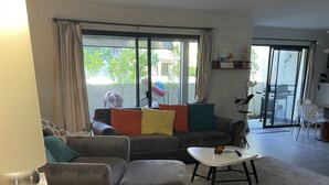 A living room with a sectional sofa, ottoman, and a small round table. There is a lamp on a cabinet, and a view of the outside through large windows. - Great Palm Springs Oasis 2 miles from downtown! (Palm Springs)