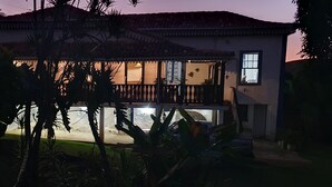 A two-story house with a balcony, surrounded by tropical plants.
