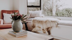 Une pièce chaleureuse avec un canapé, une fenêtre donnant sur des arbres, une table avec un vase et des livres, et un chat sur le rebord de la fenêtre.