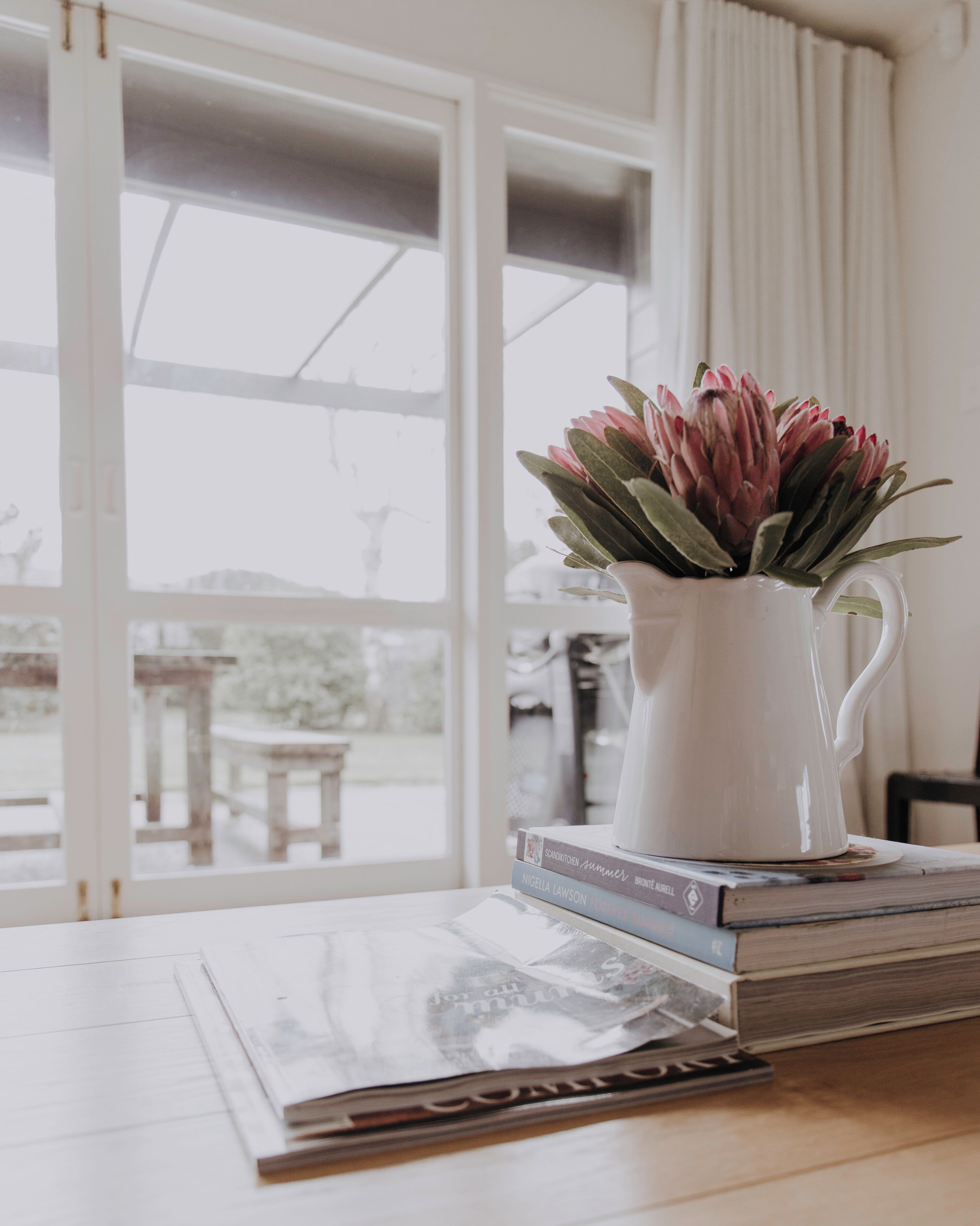 A white pitcher with pink flowers on a table, next to a window with a view of a snowy landscape.