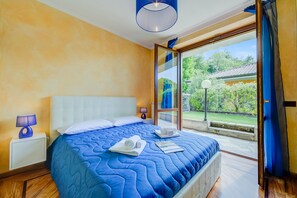 A bedroom with a large bed, a nightstand with a lamp, and a view of a garden through a glass door.