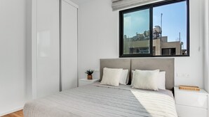 A modern bedroom with a large bed, a window with a city view, a bedside table with a plant, and a wardrobe. - Sanders Olive - Cozy 2-bdr Apt With Shared Pool (Paphos)