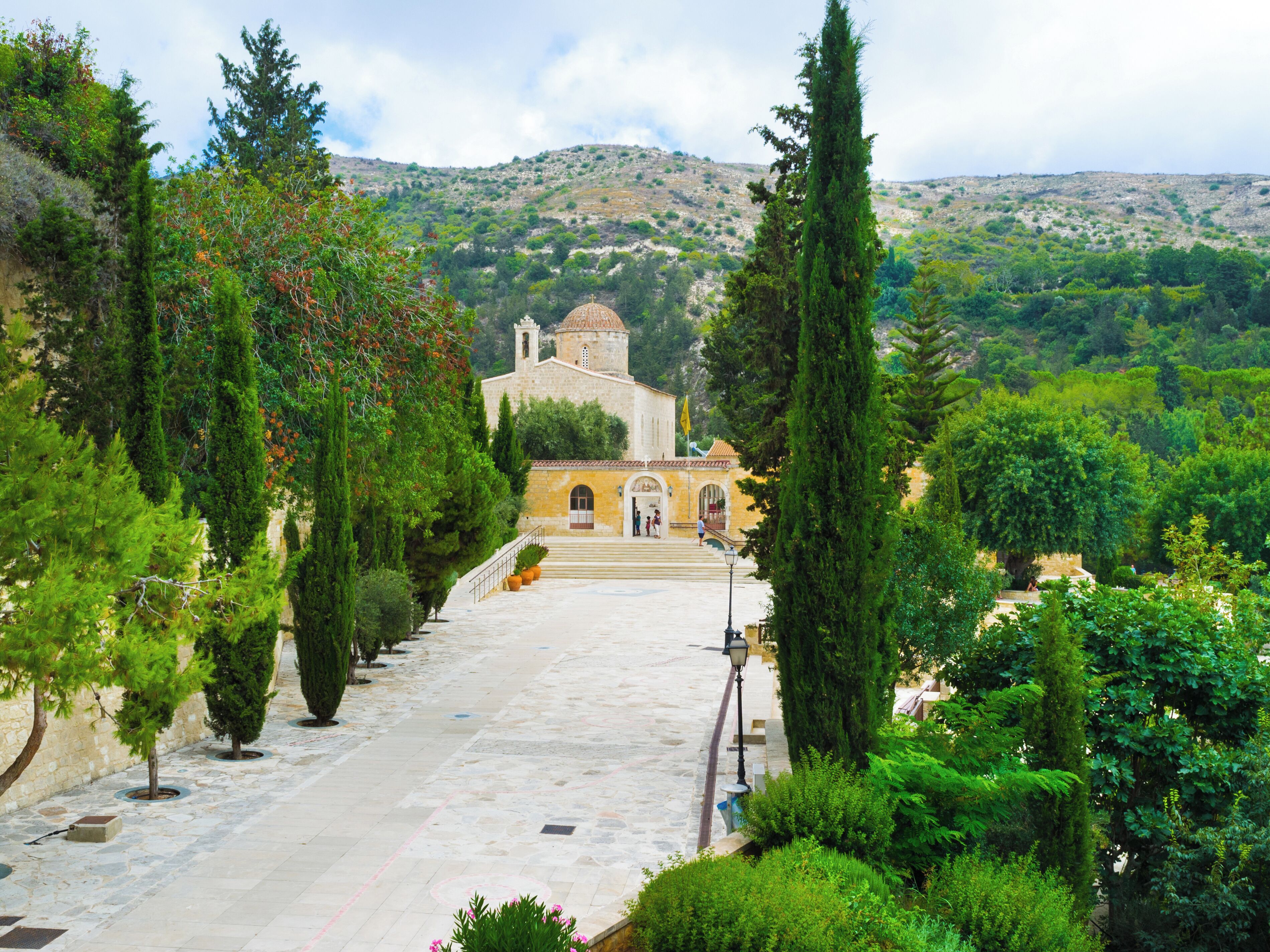 A stone pathway leading to a building with a dome and bell tower, surrounded by greenery and hills.