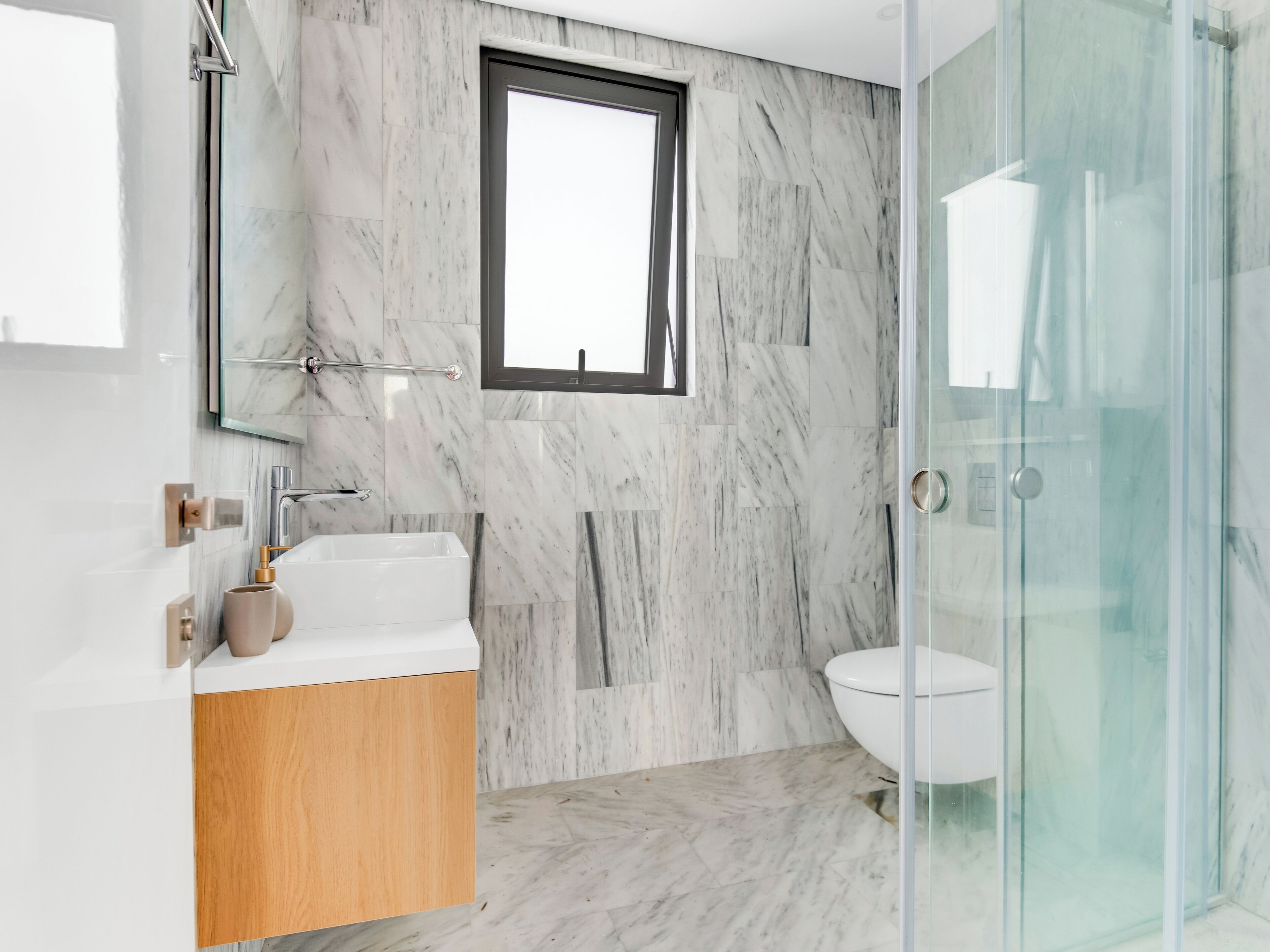 A modern bathroom with a marble wall, a window, a glass shower enclosure, and a wooden vanity.