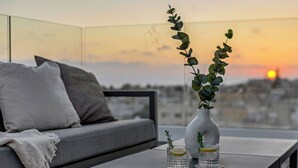 Un salón moderno con un sofá gris, un jarrón blanco con plantas verdes y dos vasos con rodajas de limón en un balcón con vista a un atardecer.