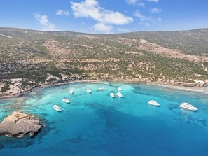 An aerial view of a coastal area with boats anchored in clear blue waters, surrounded by hilly terrain and sparse vegetation. - Sanders Olive - Cute 1-bdr Apt With Shared Pool (Paphos)