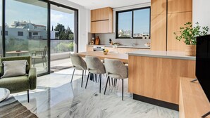 A modern kitchen with a large window, wooden cabinets, and a dining area with chairs. - Sanders Olive - Cute 1-bdr Apt With Shared Pool (Paphos)