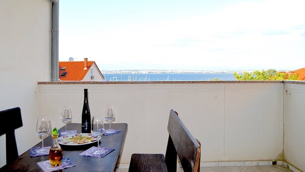 A balcony with a table set for dining, a bottle, and glasses, overlooking a coastal view with buildings in the distance. - Panoramic View Apartment Marijane (Preko)