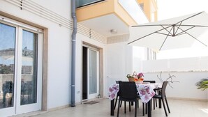 An outdoor dining area with a table set for two, a large umbrella, and a view of the cityscape through the sliding glass doors. - The House of Corals (Monopoli)
