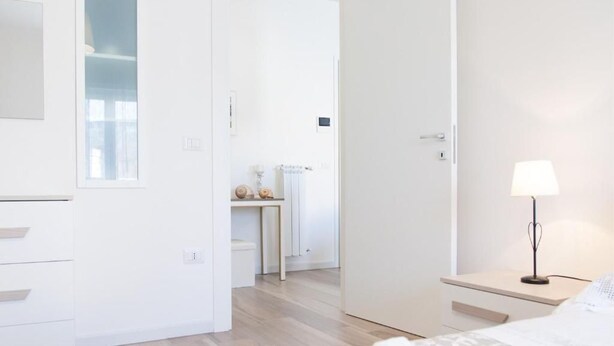 A modern hotel room with a bed, bedside table, and a lamp. There is a door leading to another room and a small table with decorative items. - The House of Corals (Monopoli)