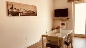 A dining area with a table and chairs, a painting of a cityscape, a television on a stand, and a window with curtains. - The House of Corals (Monopoli)