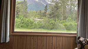 Een kamer met een groot raam dat uitkijkt op een bergachtig landschap, houten wanden en een uitzicht op bomen en een huis.